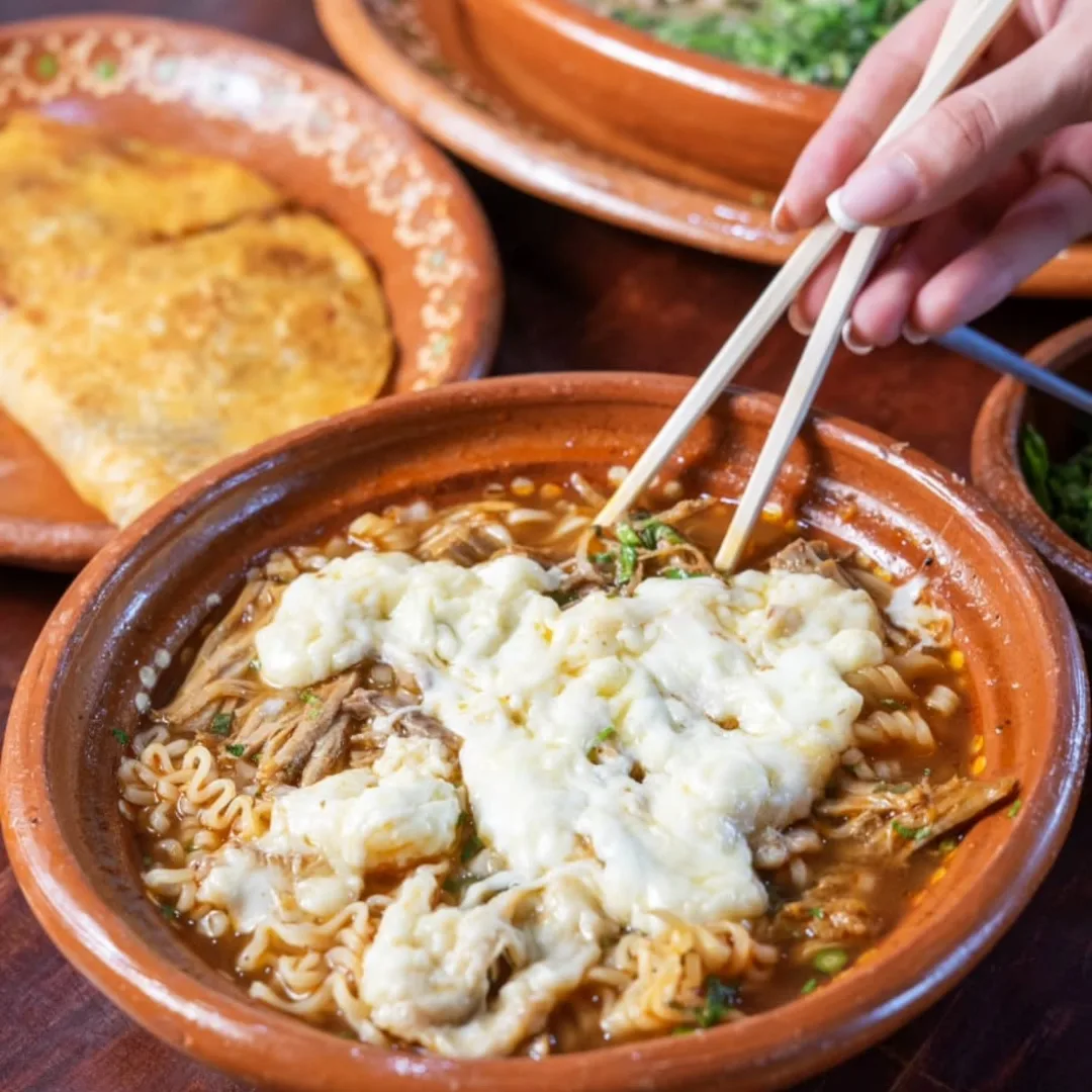 Birriamen en cazuela de barro, con los fideos levantados por los palillos y el queso derretido encima.