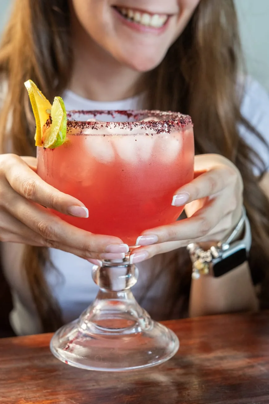 Una clienta sonríe con una copa de bebida roja escarchada con Tajín y una rodaja de naranja.