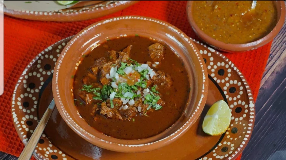 Plato de birria de res en cazuela de barro, con cilantro, cebolla y limón.